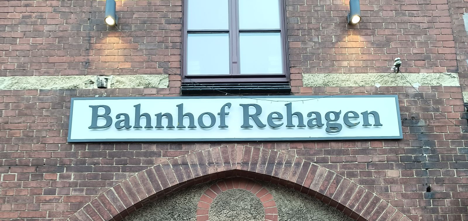 Eine Wand vom Bahnhof aus rotem Back-Stein. Auf einem Schild steht: Bahnhof Rehagen. Darunter sind zwei hohe Fenster.