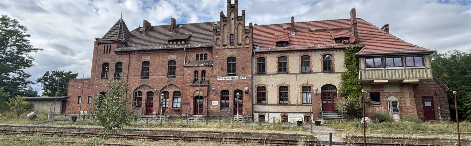 Der Bahnhof Rehagen. Das Haus ist aus rotem Back-Stein. Die Sonne scheint. Vor dem Bahnhof sind alte Gleise. Links stehen Bäume.